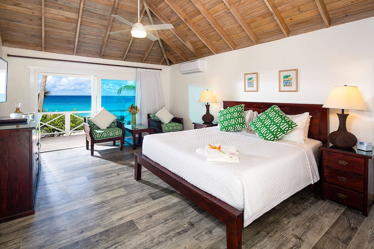 A bedroom with a wooden ceiling, a bed with white bedding, green patterned pillows, a nightstand with a lamp, and a view of the ocean.