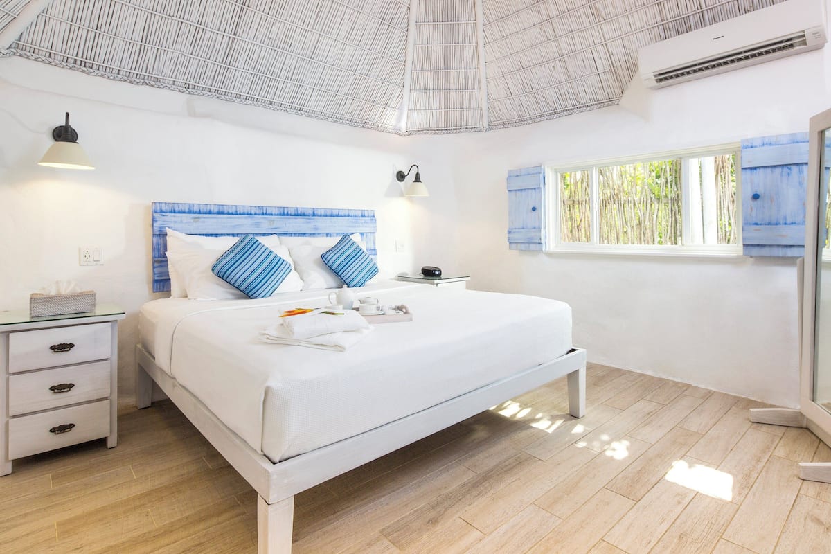 A modern bedroom with a large bed, bedside tables, a window with bamboo shutters, and a ceiling with a unique woven design.