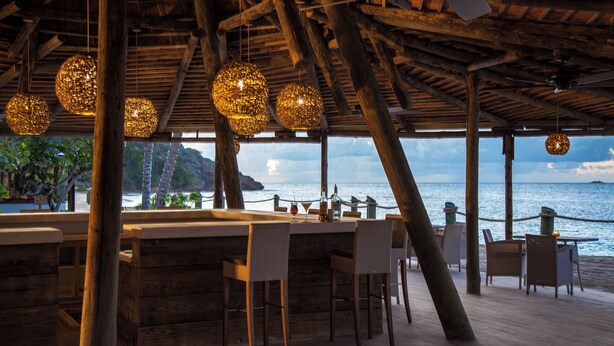Un espace bar en bois, agrémenté de luminaires sphériques suspendus, qui offre une vue sur une plage, l’océan et la côte lointaine.