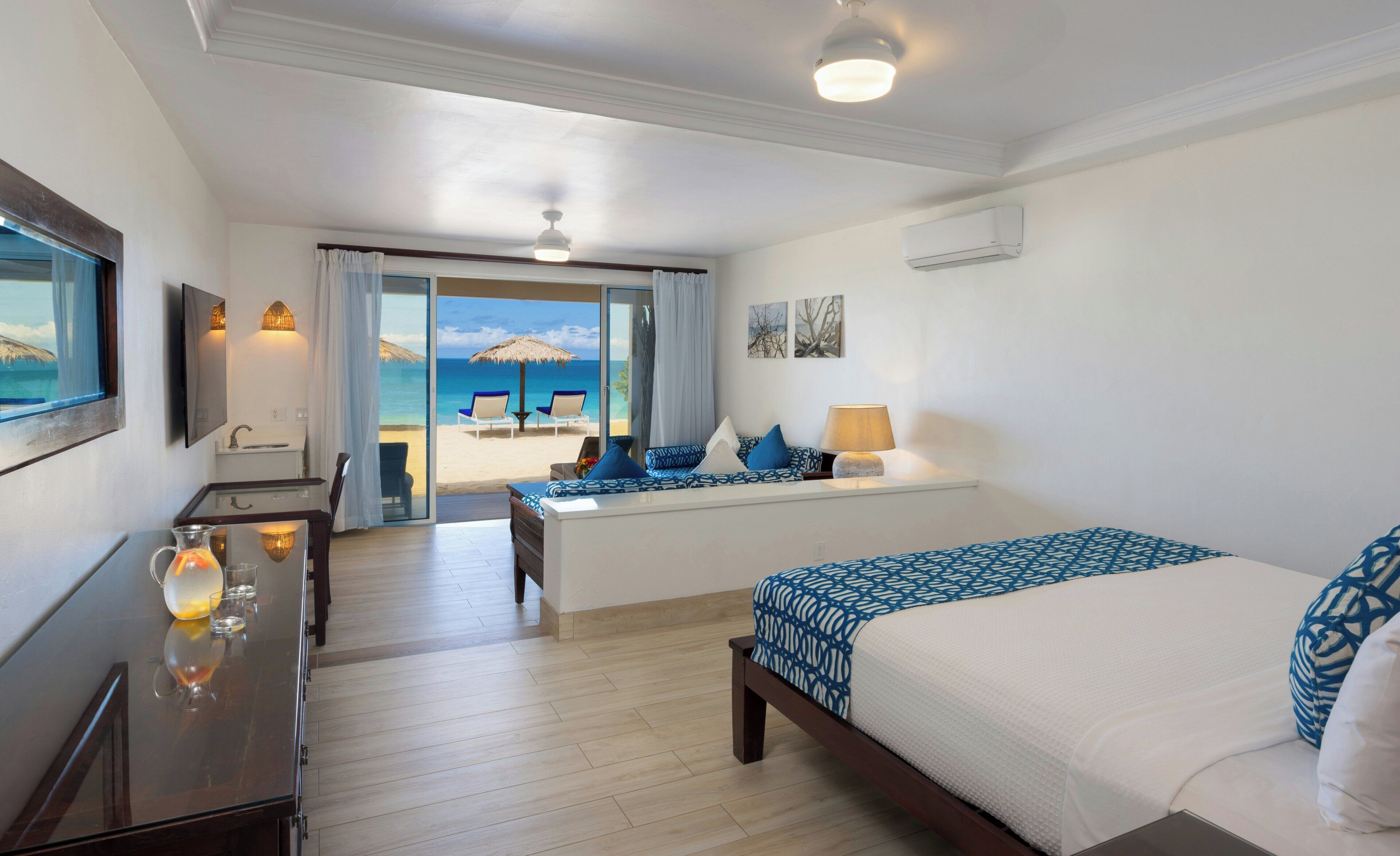A hotel room with a bed, a desk, a chair, a small table with a pitcher and glasses, and a view of the beach through a sliding door.