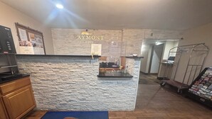 Zona de recepción con pared de piedra, mostrador de recepción, felpudo azul con el nombre del hotel y un carrito con revistas.