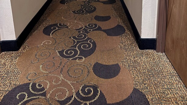 Un pasillo de hotel con pisos alfombrados, puertas a ambos lados y un largo corredor que conduce a una habitación luminosa al final.