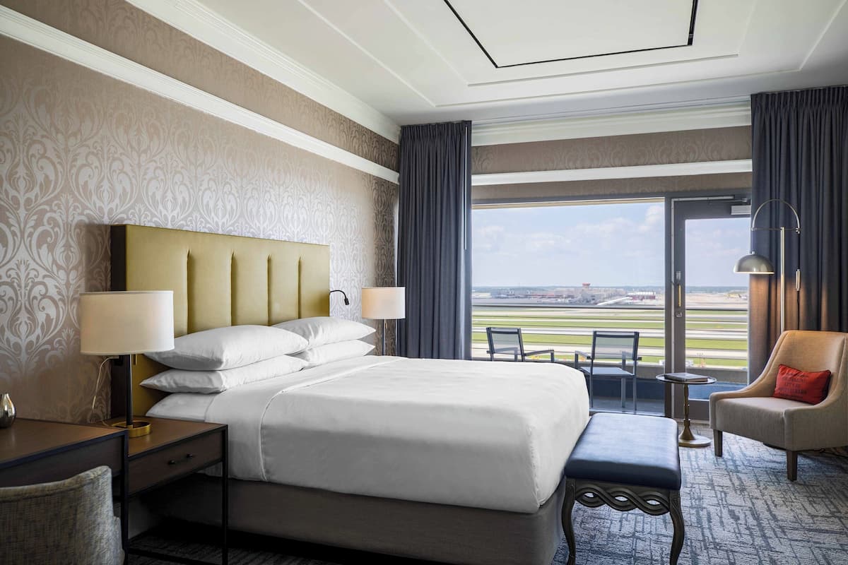 A hotel room with a large bed, two bedside tables with lamps, a chair, a small table, and a view of the airport.