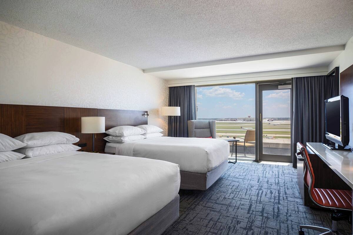 A hotel room with two beds, a desk with a TV, a chair, and a view of the airport.