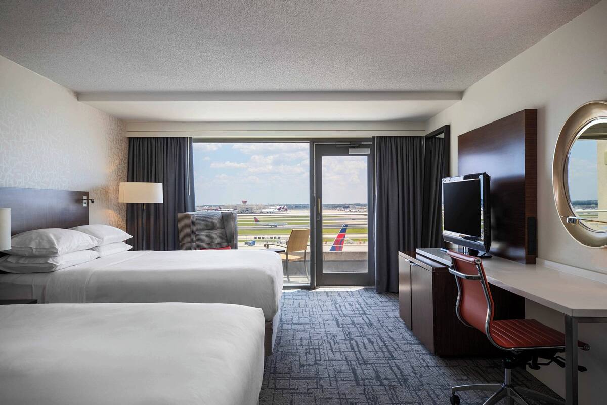 A hotel room with two beds, a desk with a chair, a television, and a large window offering a view of an airport.