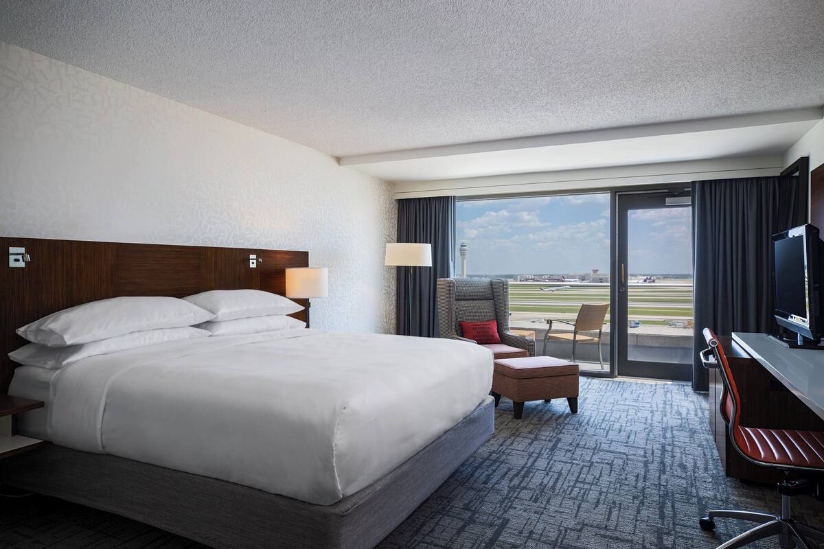 A hotel room with a large bed, a desk with a TV, a chair, and a view of the airport.