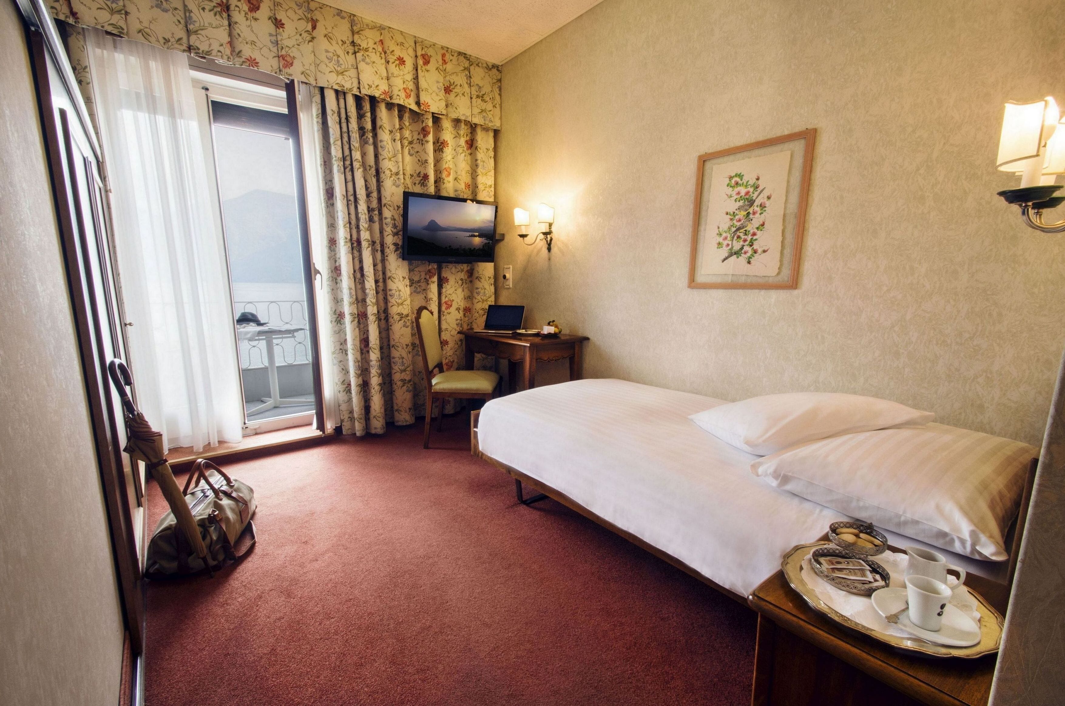 A hotel room with a bed, a chair, a desk with a laptop, a TV, and a tray with a cup and saucer.