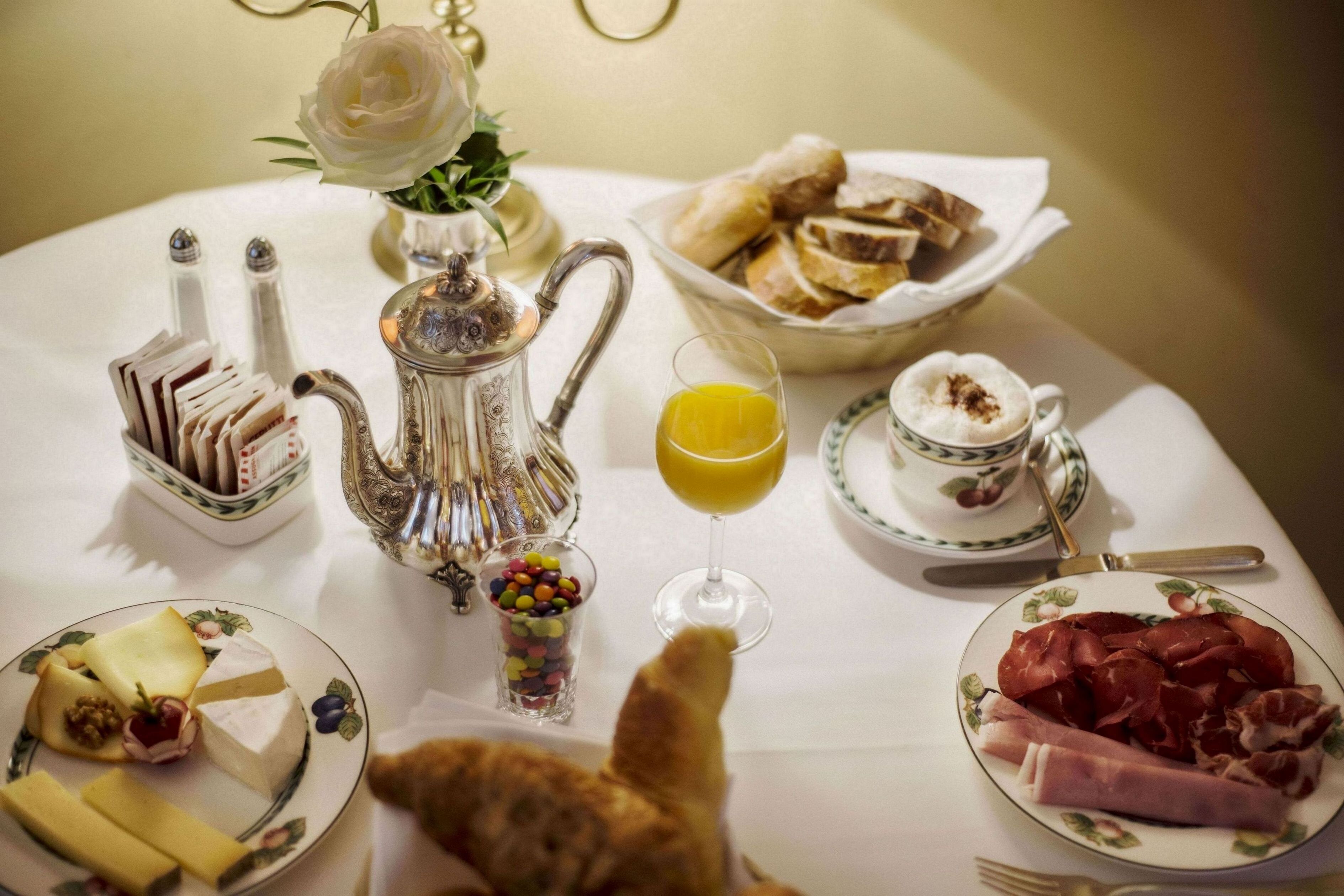 A breakfast table with various food items, a teapot, a glass of orange juice, and a cat.