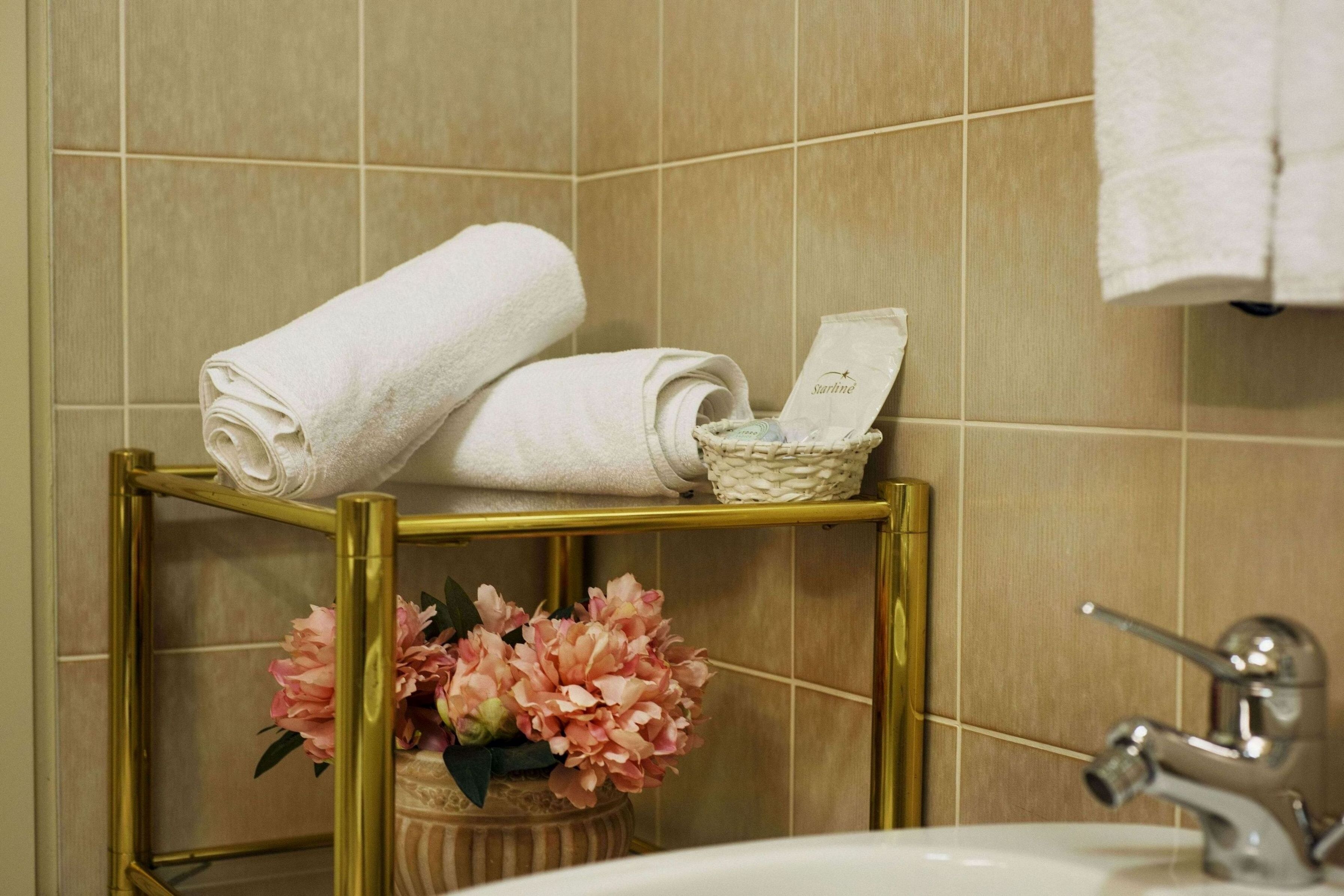 Ein Badezimmer mit beigen Fliesen, einem goldfarbenen Waschtisch und einem weißen Waschbecken, sowie einer dekorativen Vase mit rosa Blumen.