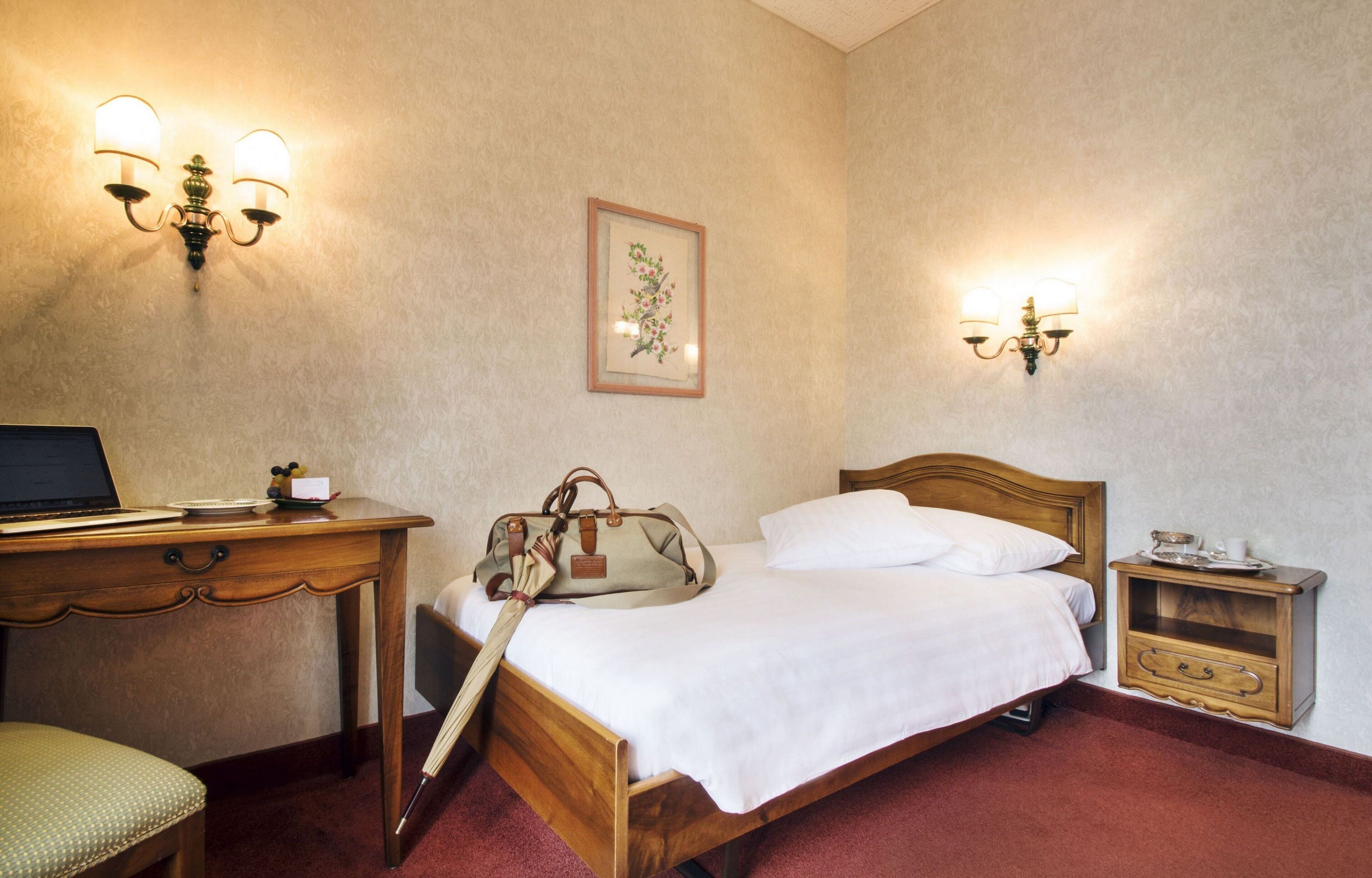 Una habitación de hotel con una cama de madera, un escritorio con una computadora portátil, una silla, una mesita de noche con una lámpara y un cuadro en la pared.