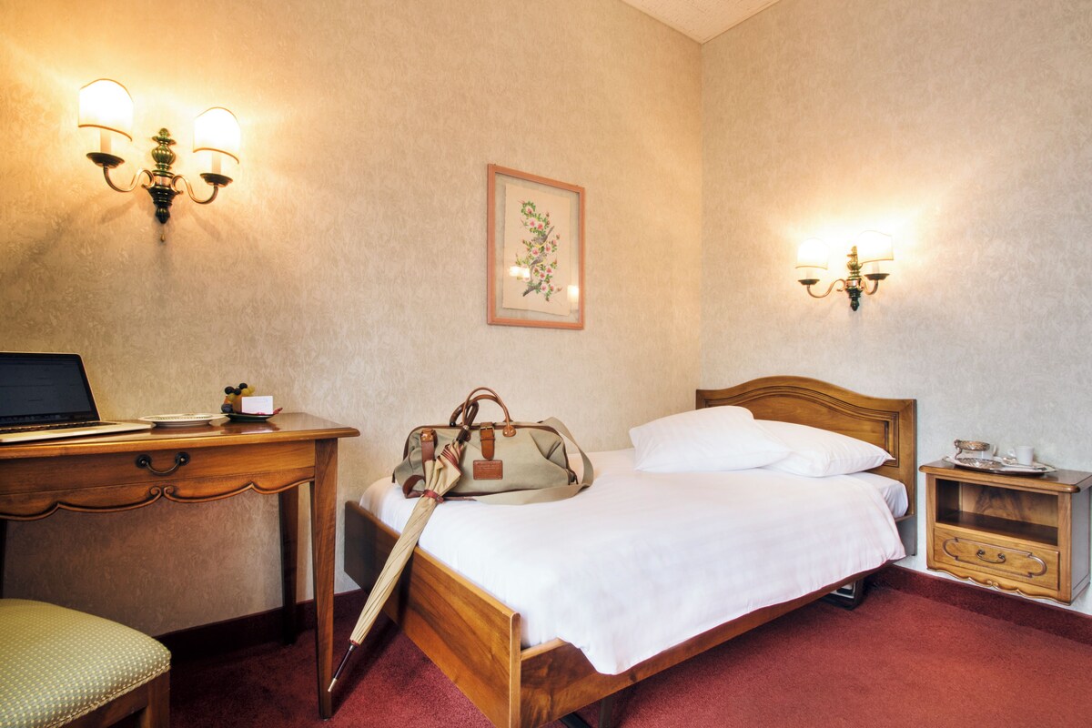 Une chambre d’hôtel avec un lit en bois, un bureau avec un ordinateur portable, une chaise, une table de chevet avec une tasse et des appliques murales.