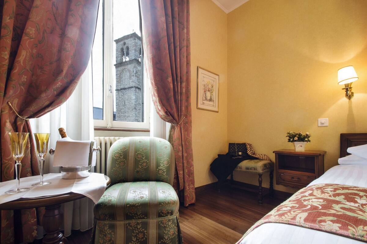 Une chambre d’hôtel avec un lit, une chaise, une table avec des verres, une lampe et une fenêtre avec des rideaux.