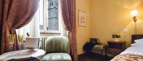 A hotel room with a bed, a chair, a table with glasses, a lamp, and a window with curtains.