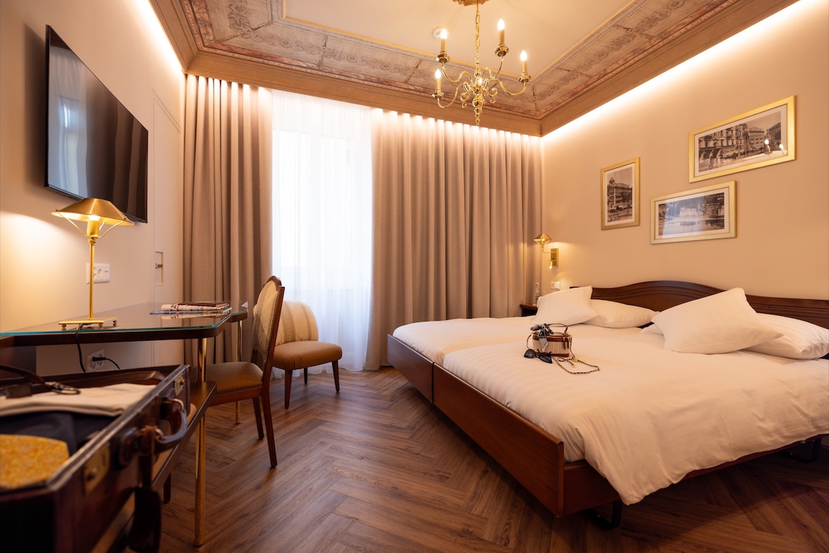 A hotel room with a bed, a desk with a lamp, a chair, a TV, and framed pictures on the wall.