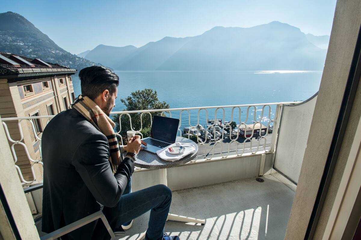 Un homme est assis sur un balcon, devant un ordinateur portable, et il a devant lui une vue sur un lac et des montagnes.