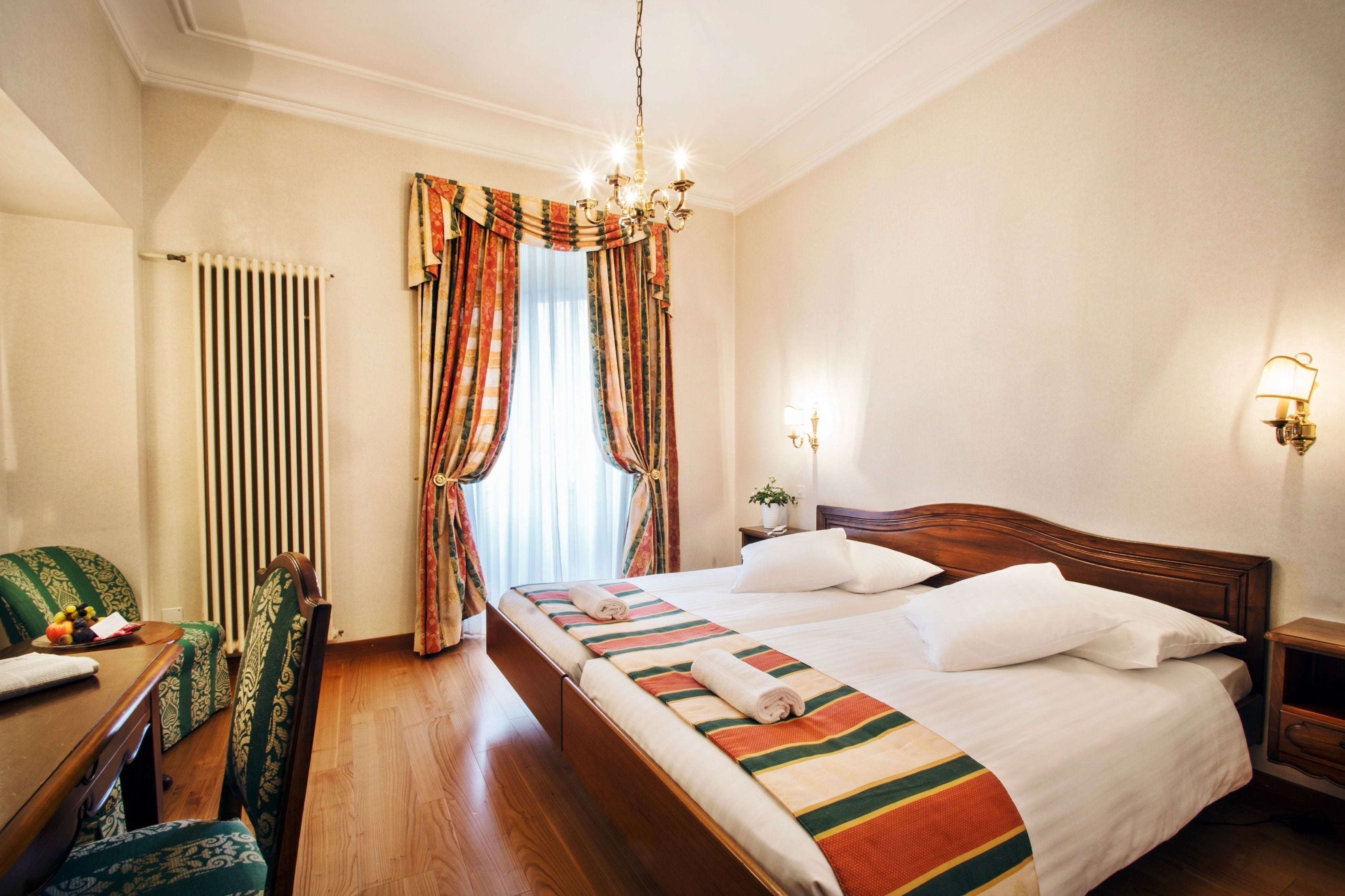 A hotel room with a large bed, a wooden headboard, a bedside table with a lamp, a chair, and a radiator.