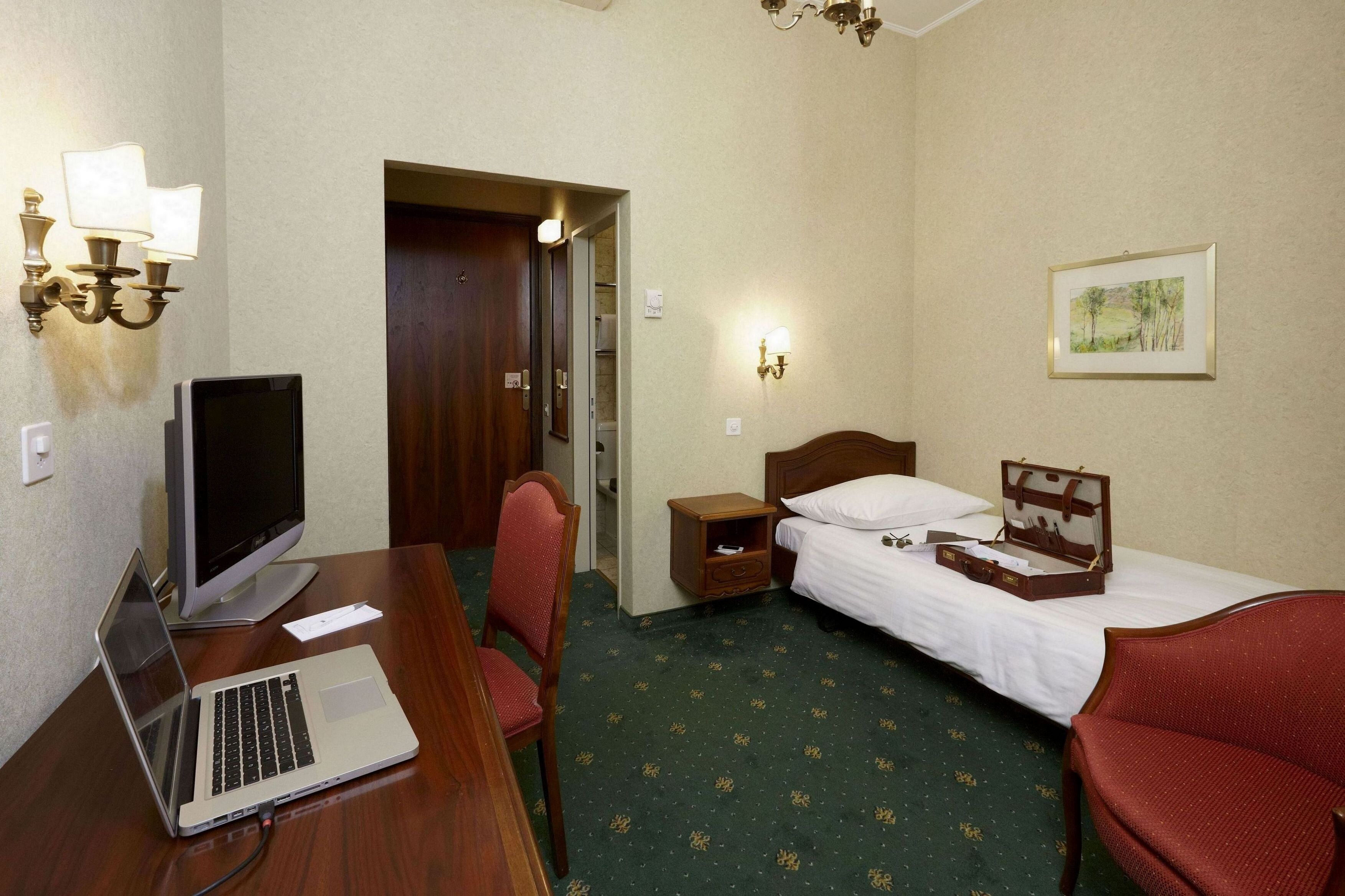 Une chambre d’hôtel comprenant un lit, un bureau avec un ordinateur portable, une chaise, une table de chevet et une lampe fixée au mur.