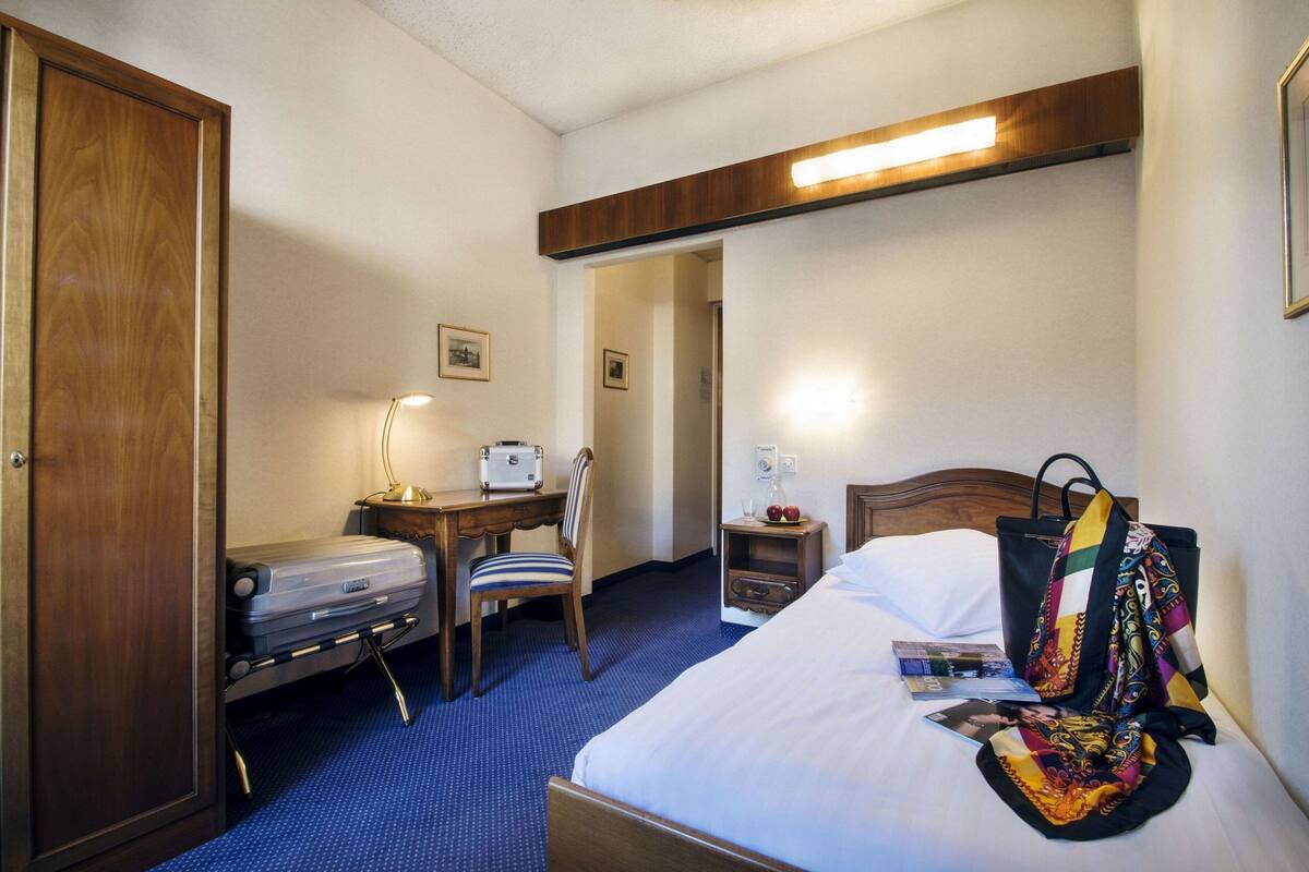 Une chambre d’hôtel comprenant un lit, un bureau avec une valise, une chaise, une table de chevet et une armoire en bois.