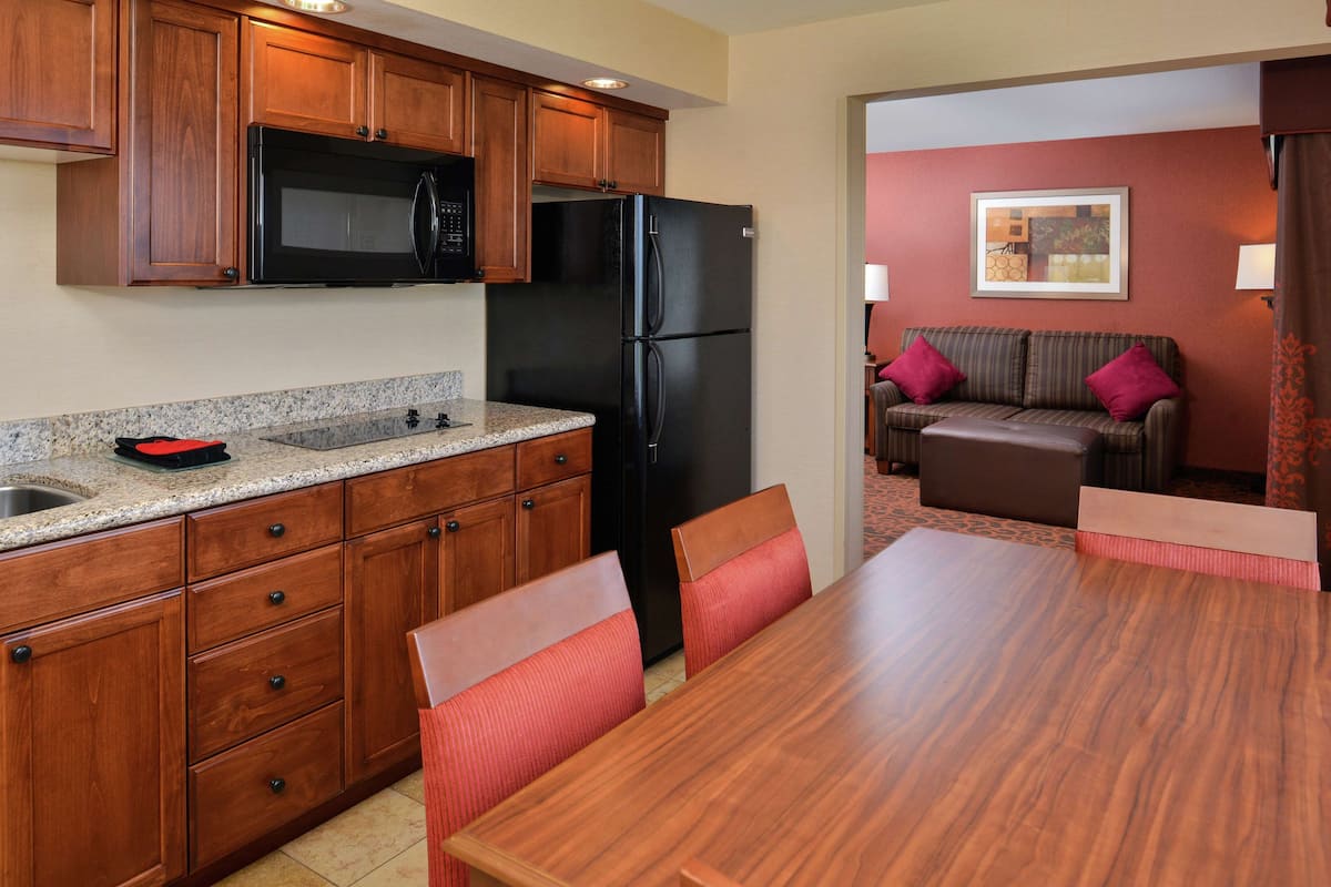 A kitchen with wooden cabinets, a black refrigerator, and a granite countertop. There is a dining table with chairs and a sofa in the adjacent living area.