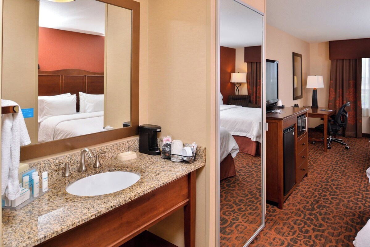 A hotel room with a granite countertop sink, a mirror, a bed with white linens, a desk with a chair, and a patterned carpet.