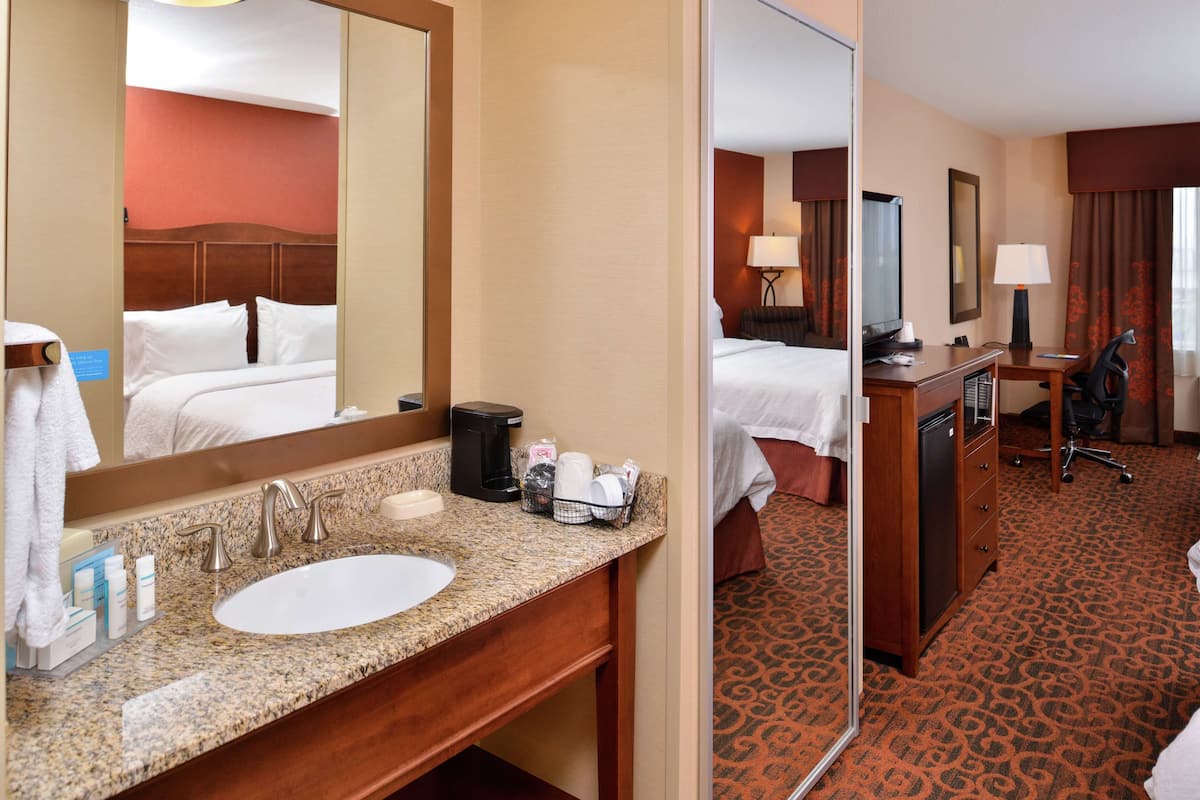A hotel room with a large bed, a bathroom with a sink and mirror, a desk with a chair, and a carpeted floor.