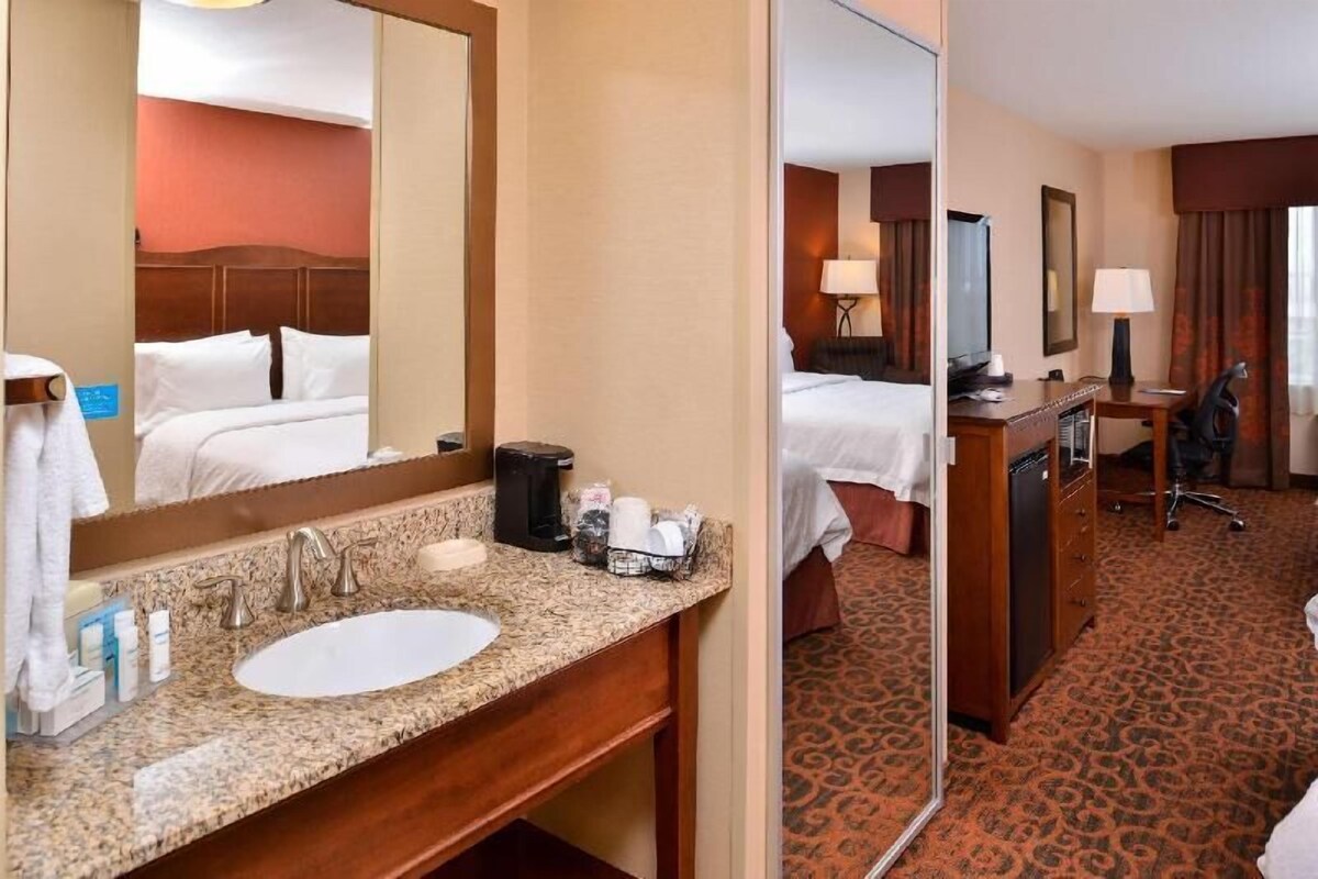 A hotel room with a bed, a desk, a chair, a desk lamp, a television, a mirror, a sink, and toiletries.