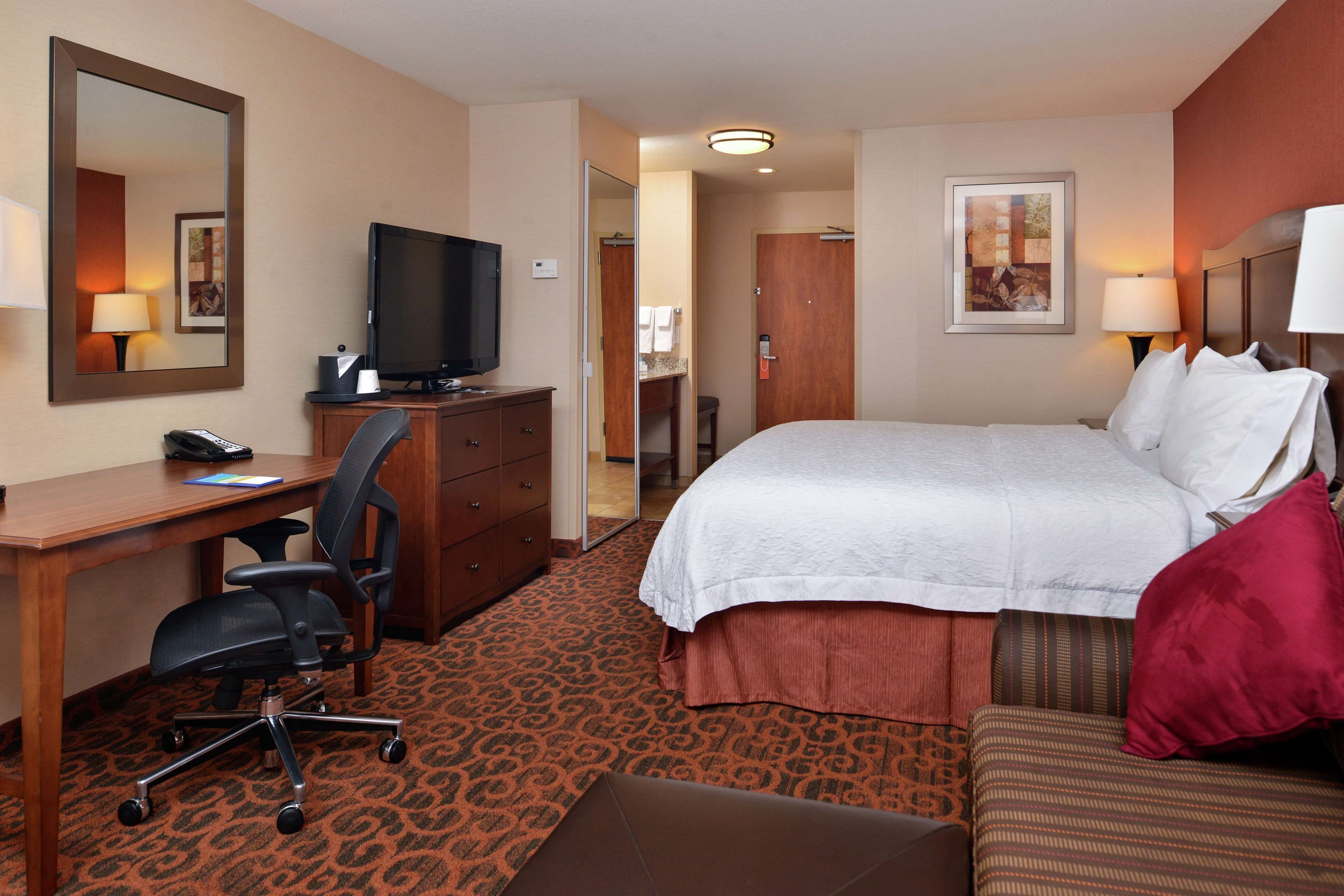 A hotel room with a bed, a desk with a chair, a television, and a bathroom visible through an open door.