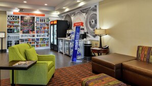 A lobby with a green armchair, a brown sofa, a patterned ottoman, a small table with a magazine, a refrigerator, a counter with a 'Smartwater' sign, and shelves with various items. - Hampton Inn Idaho Falls/Airport (Idaho Falls)
