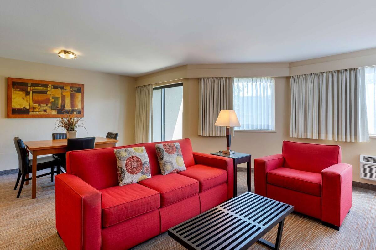 A hotel room with a red sofa, a coffee table, a dining table with chairs, a lamp, and a painting on the wall.