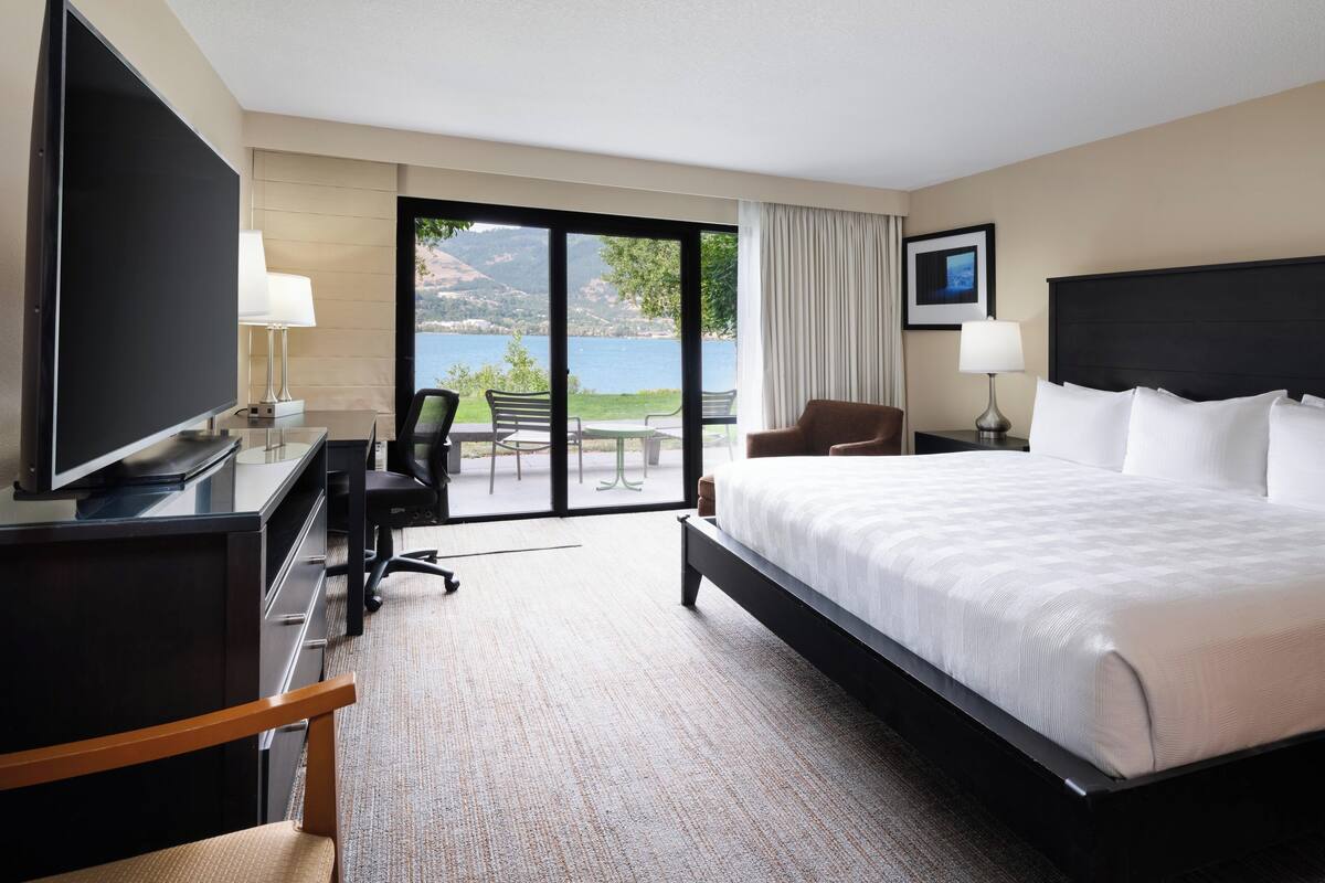 A hotel room with a large bed, a desk with a TV, a chair, a desk lamp, and a view of the outdoors.