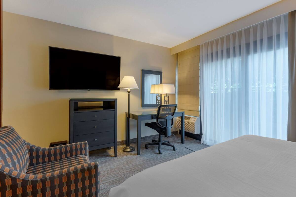 A hotel room with a bed, a desk with a chair, a television, a dresser, and a window with curtains.