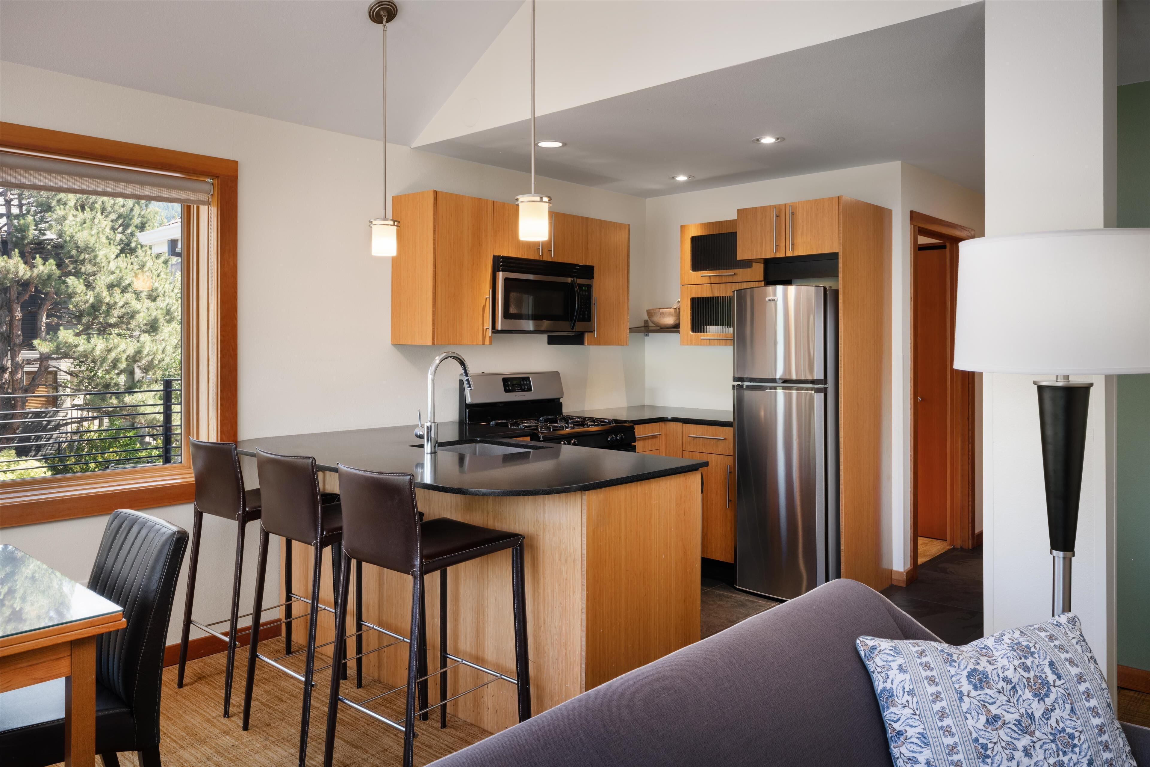 A modern kitchen with wooden cabinets, stainless steel appliances, a central island with bar stools, and a view of the outdoors through a large window.