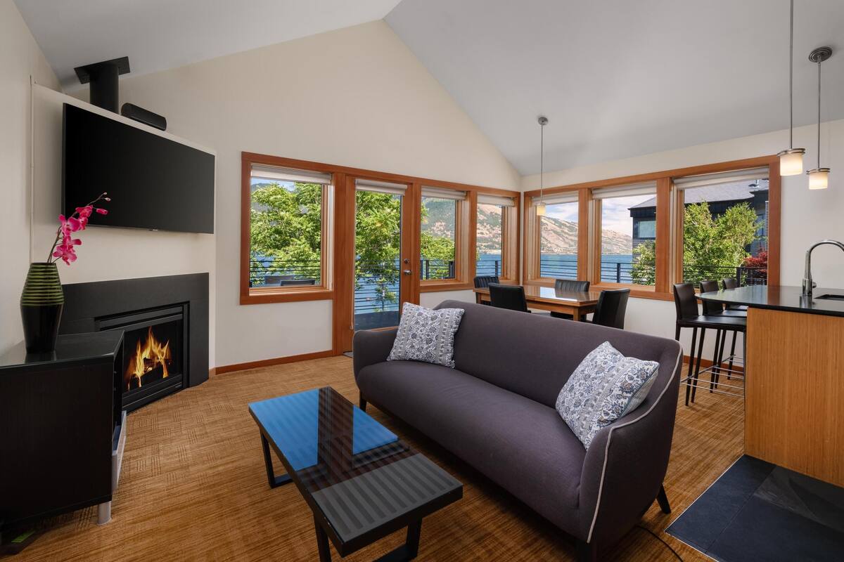 A modern living room with a fireplace, a sofa, a dining area, and large windows offering a view of the outdoors.