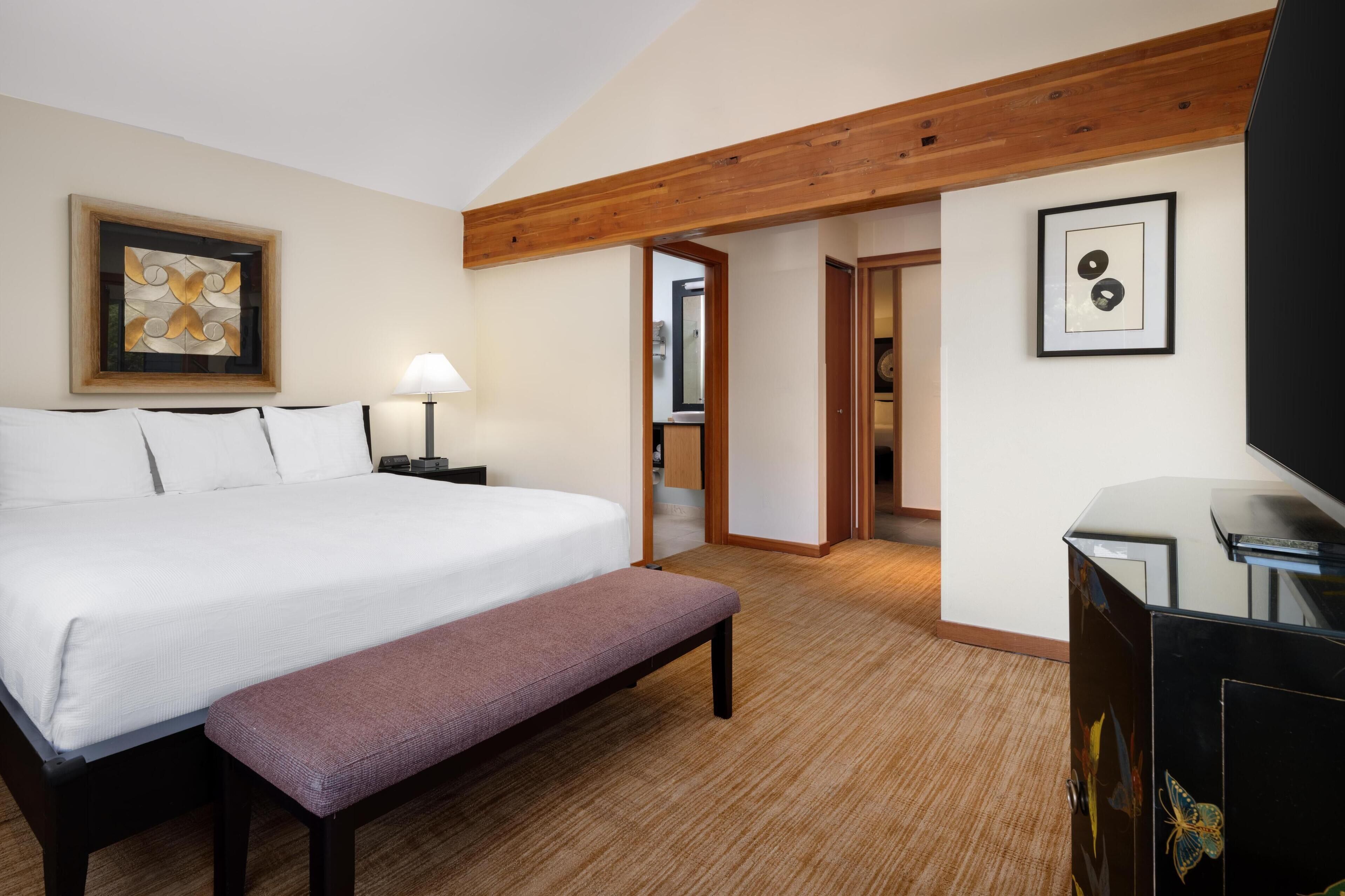 Habitación de hotel con una cama grande, un banco, un escritorio con televisor, una lámpara y obras de arte en las paredes.