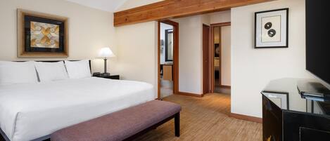 A hotel room with a large bed, a bench, a desk with a TV, a lamp, and artwork on the walls.