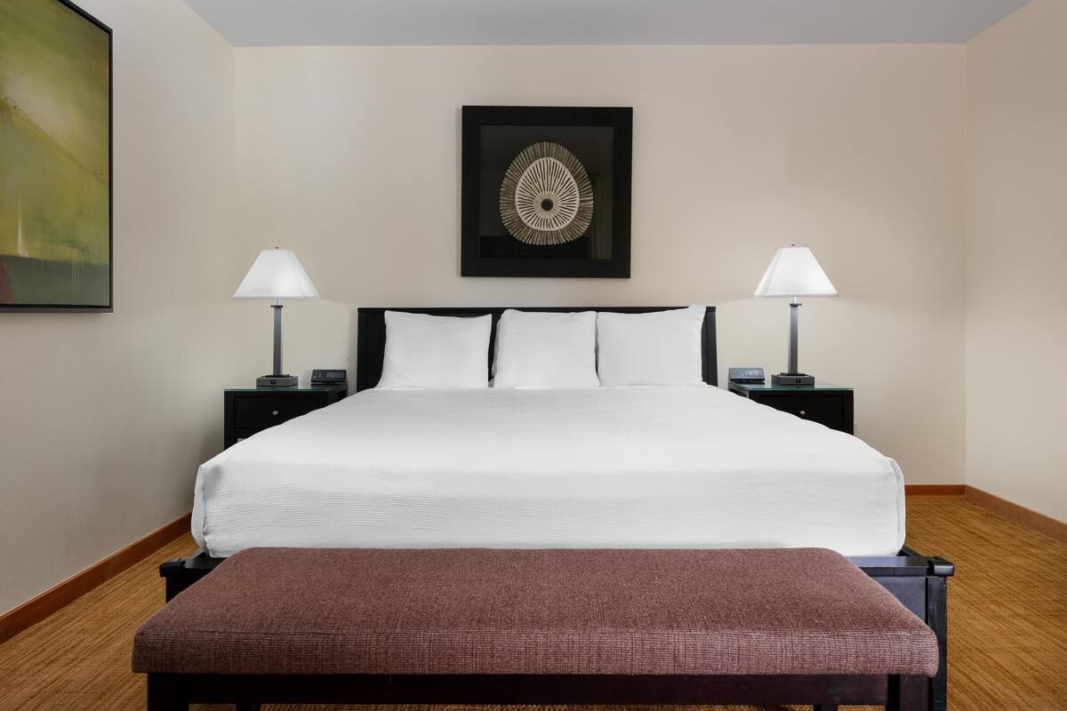 A hotel room with a large bed, two bedside tables with lamps, a bench at the foot of the bed, and a framed artwork on the wall.