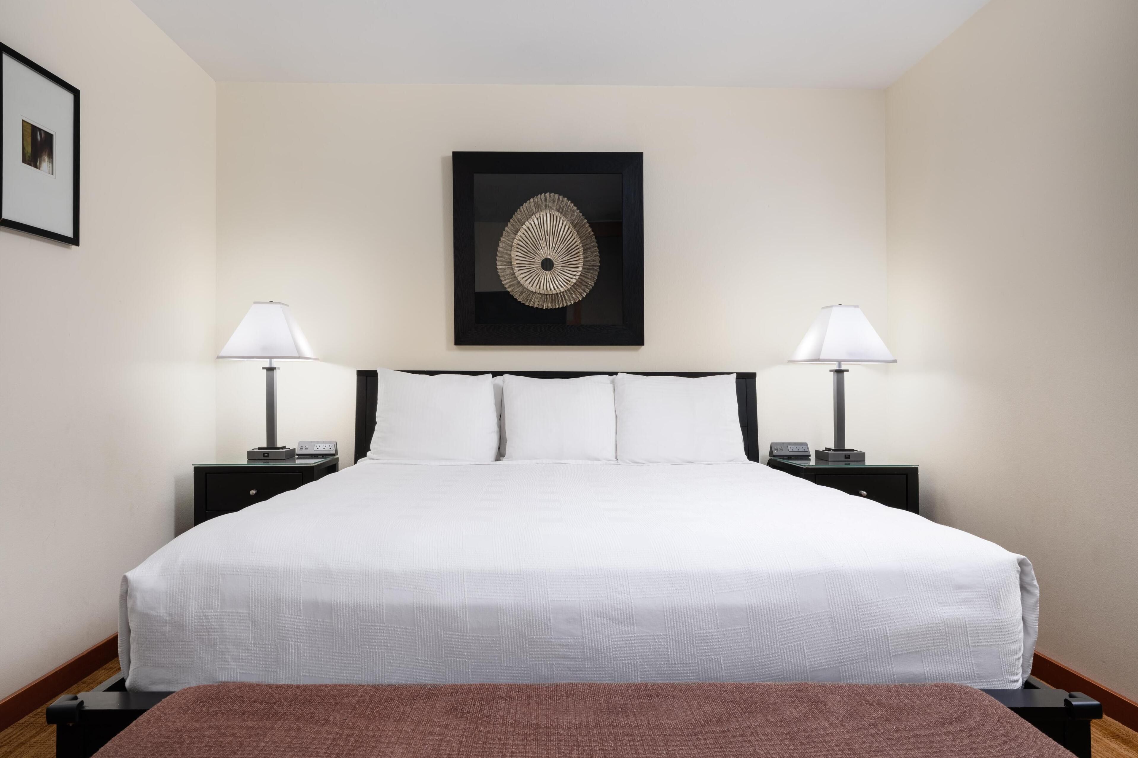 A neatly made bed with white linens, two bedside tables with lamps, and a framed artwork above the bed.