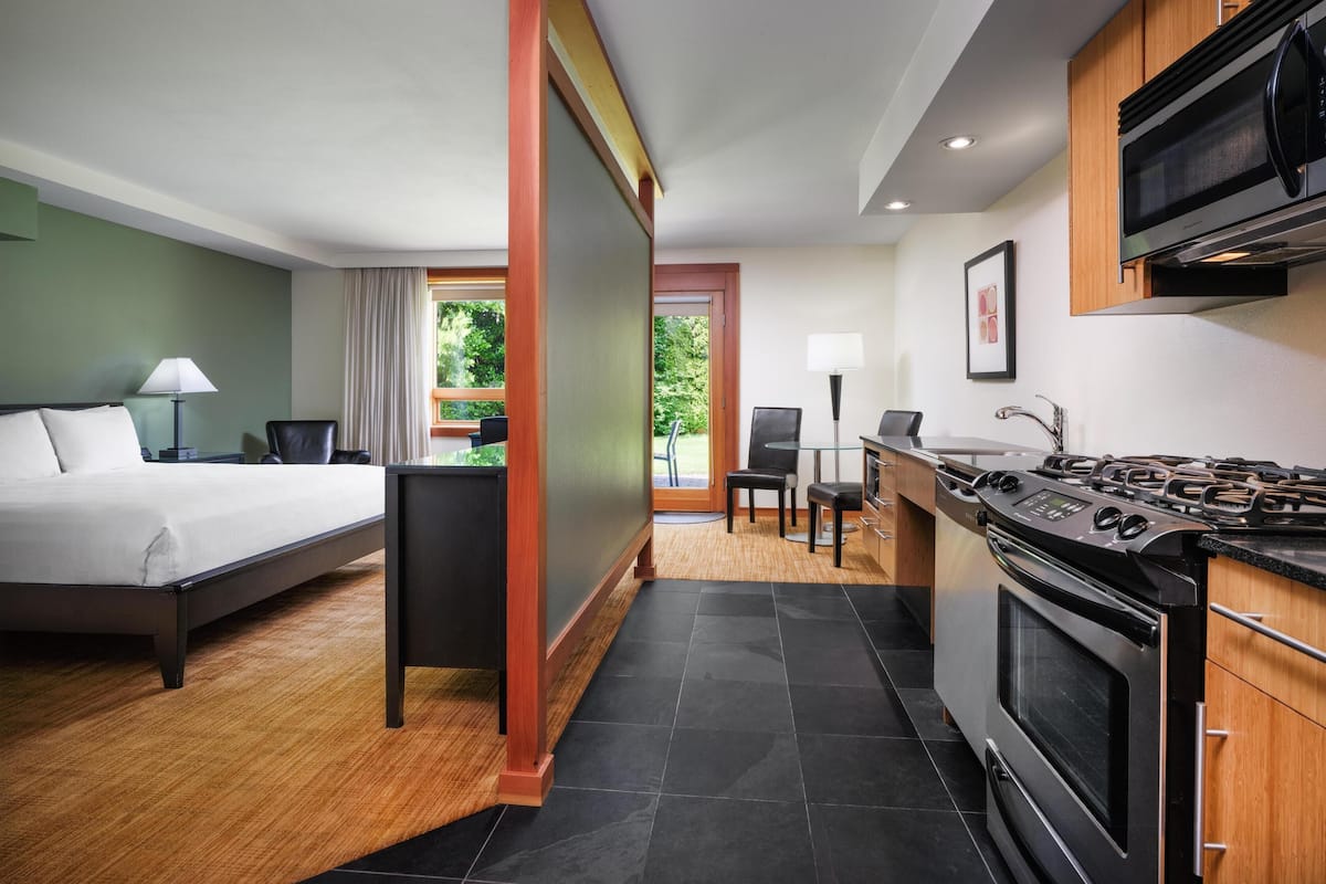 A hotel room with a bed, a desk, a chair, a kitchenette with a stove, and a view of greenery outside.