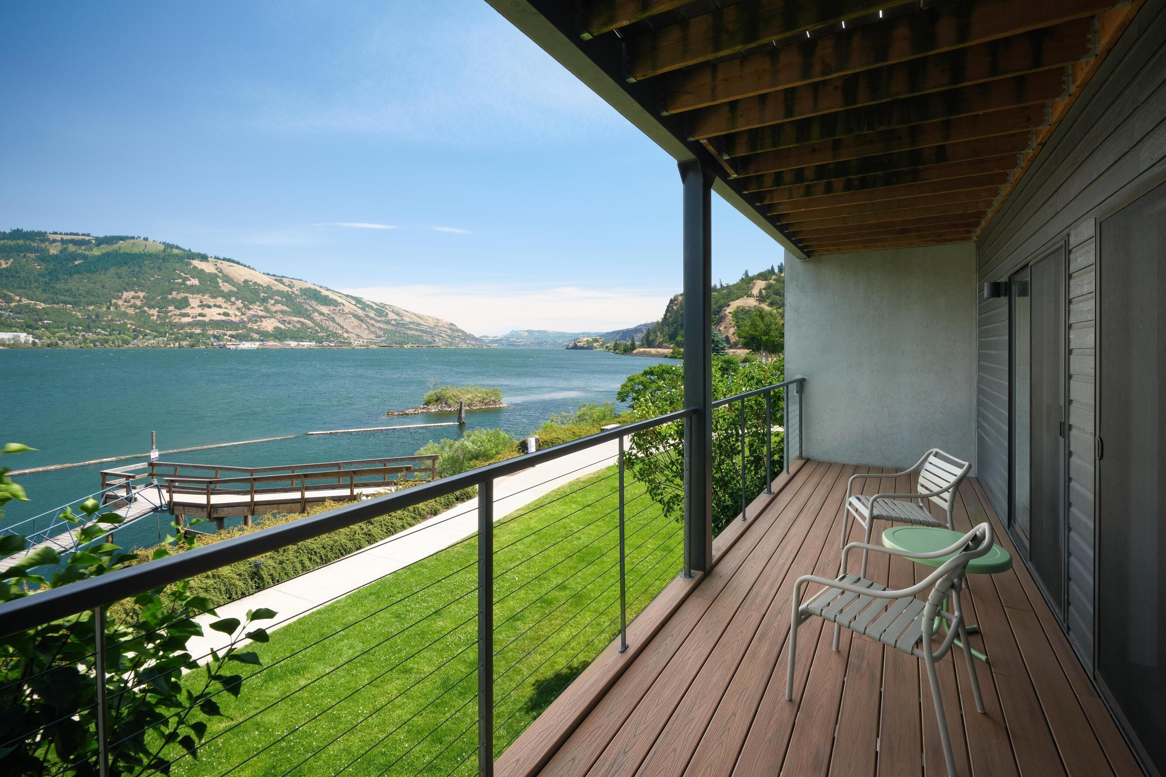 A balcony with a view of a lake and hills, featuring two chairs and a table.