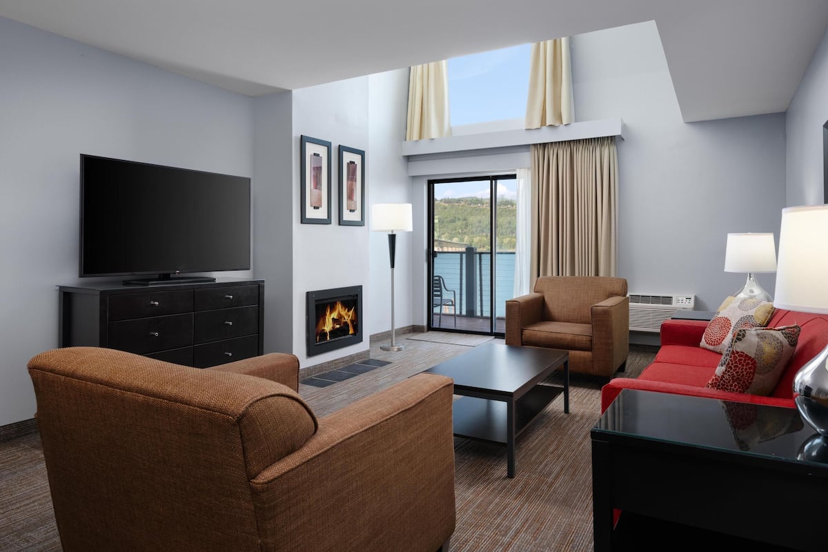 A modern hotel room with a flat-screen TV, a fireplace, a red sofa, a brown armchair, and a glass table.