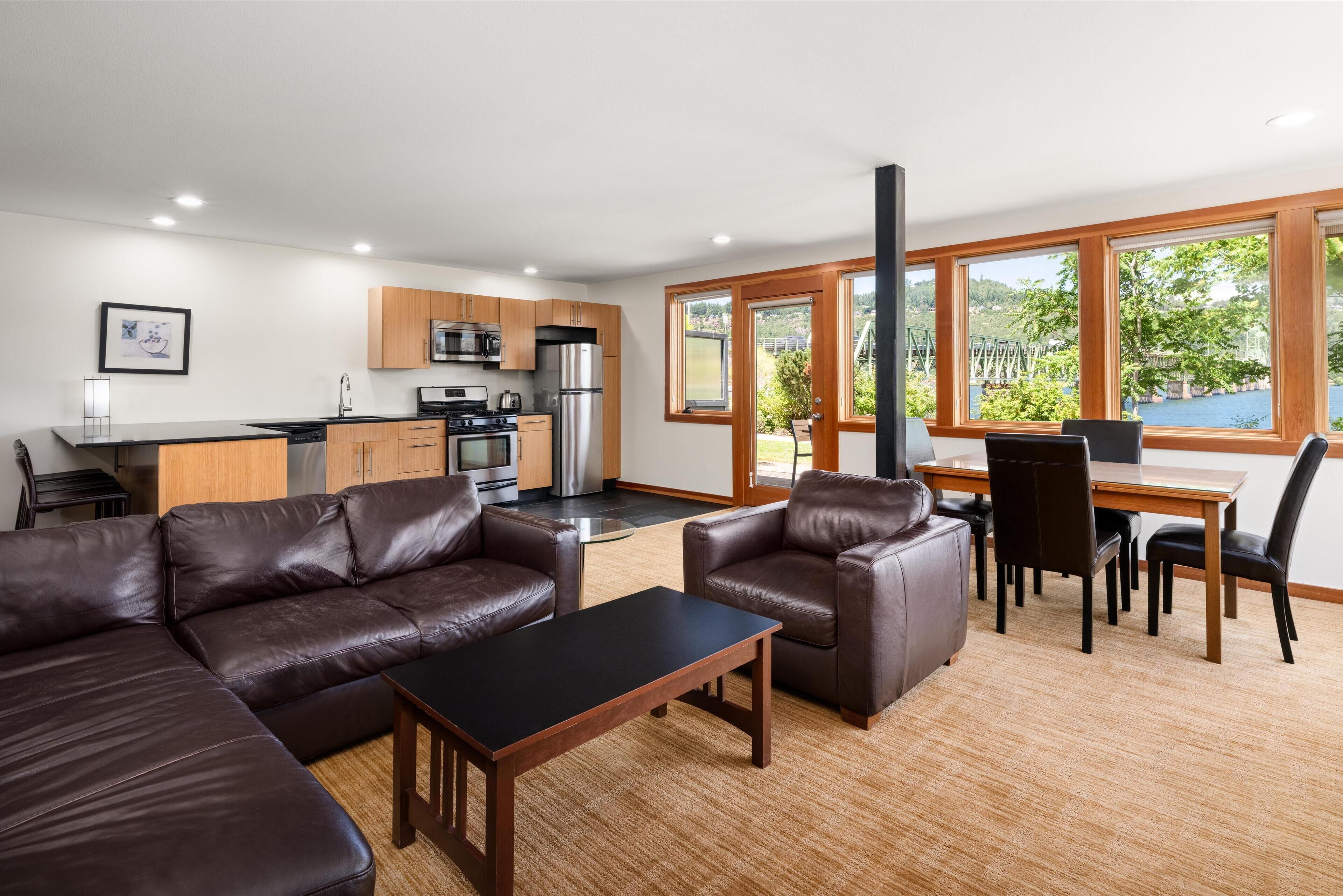 A modern kitchen with stainless steel appliances, a dining area with a wooden table and chairs, and a large window offering a view of greenery and a body of water.
