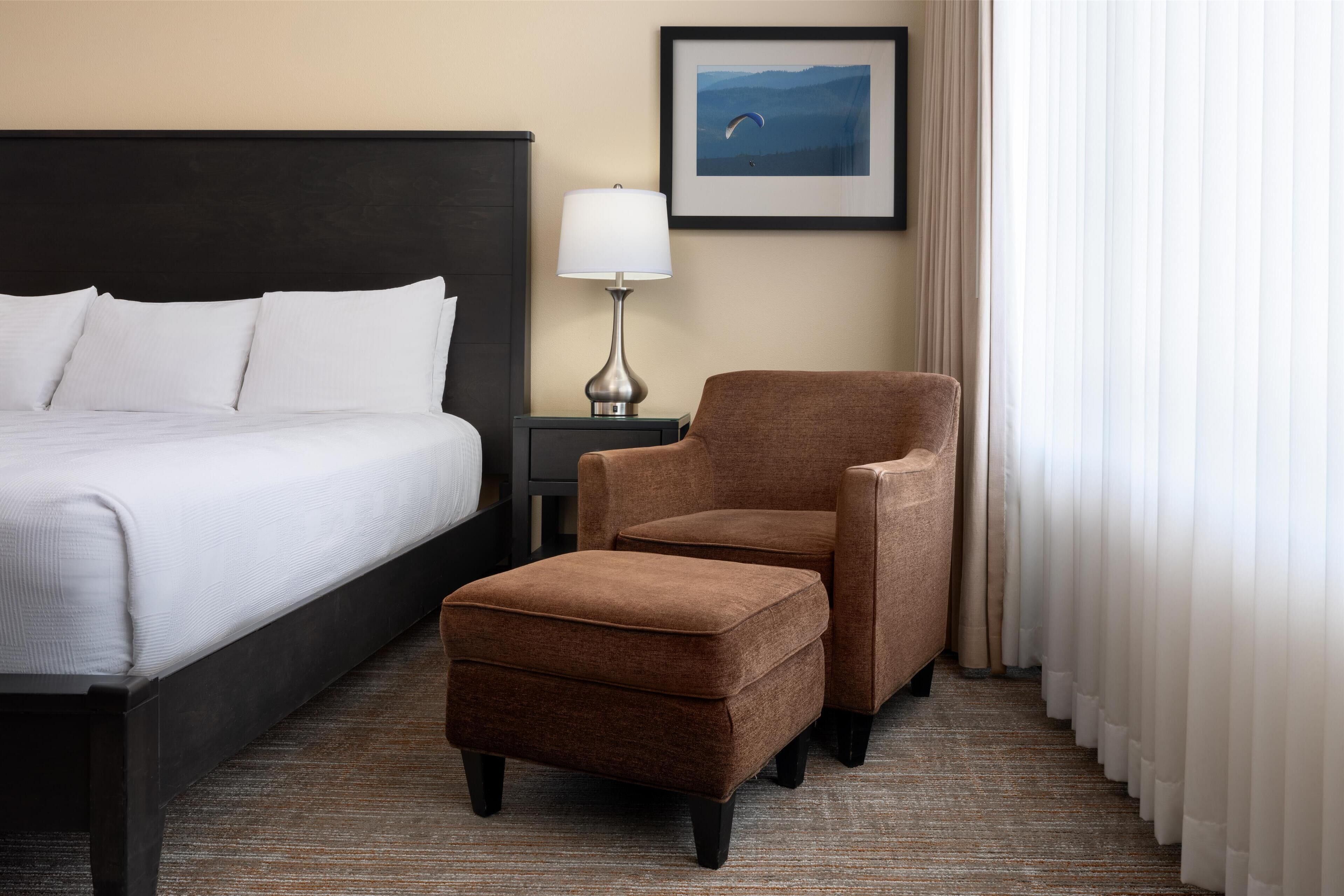 A hotel room with a bed, a brown armchair, a small ottoman, a nightstand with a lamp, and a framed picture on the wall.