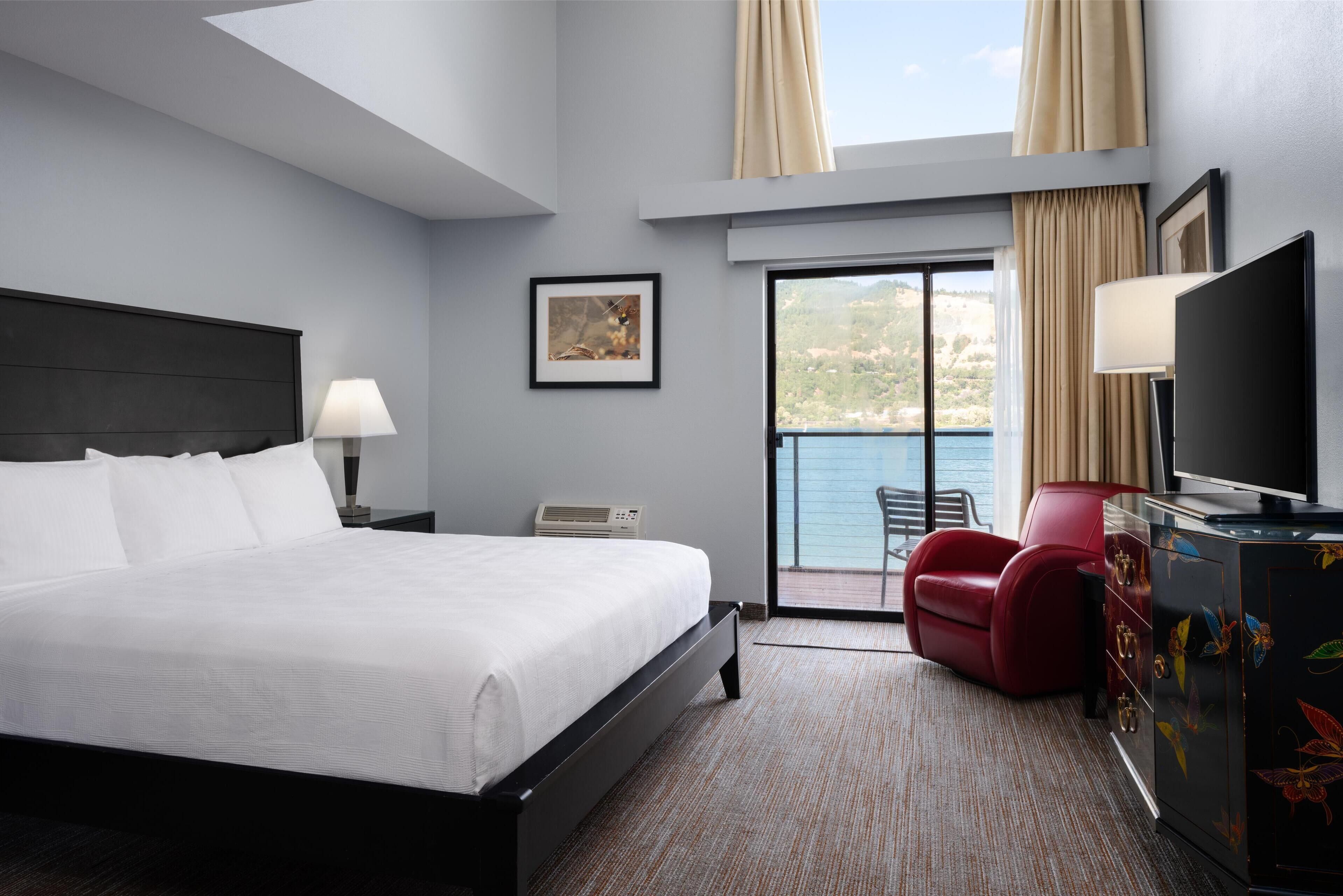 Une chambre d’hôtel avec un grand lit, un fauteuil rouge, une télévision et un balcon donnant sur la mer.