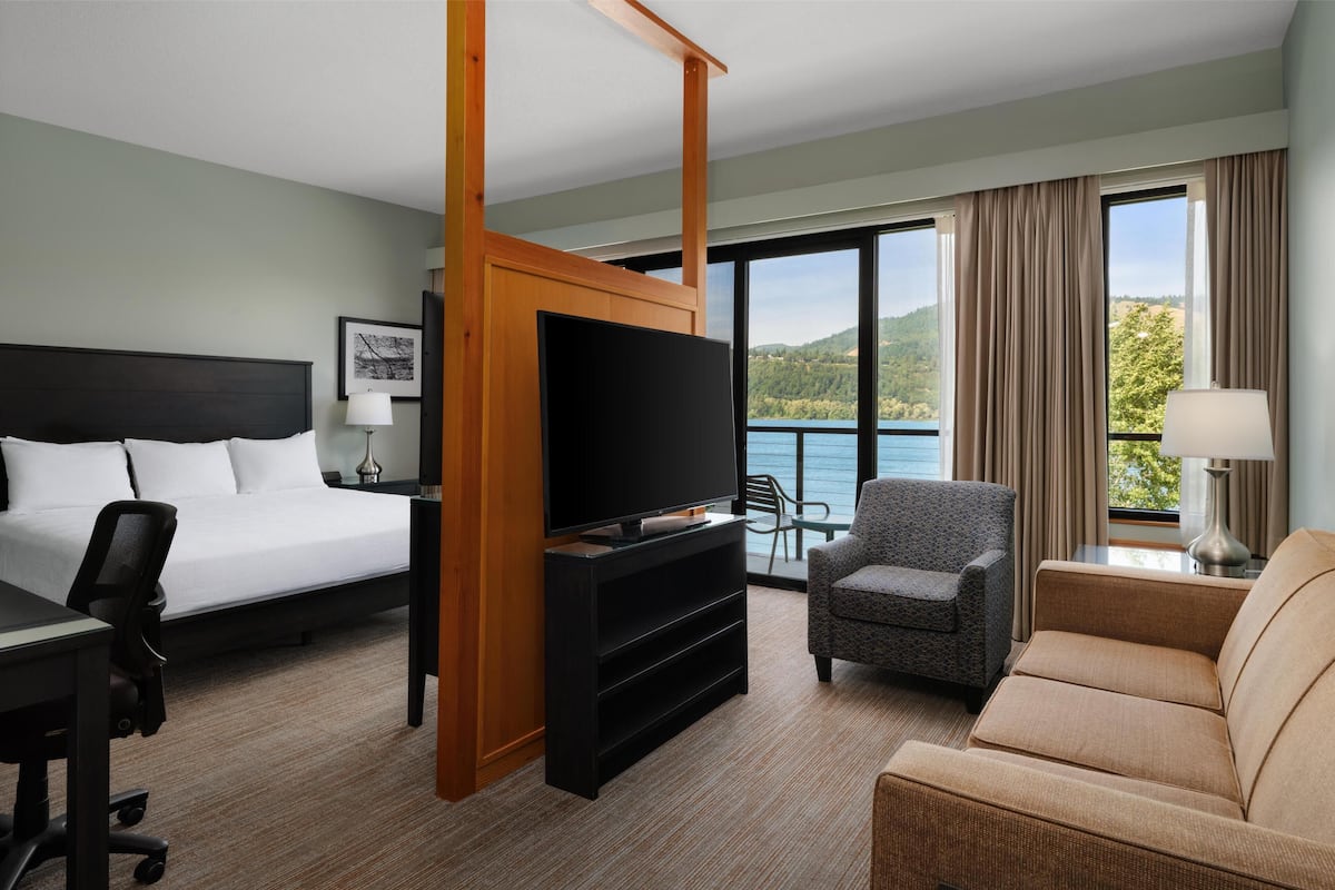 A hotel room with a bed, a TV, a desk, a chair, a sofa, and a view of a lake.