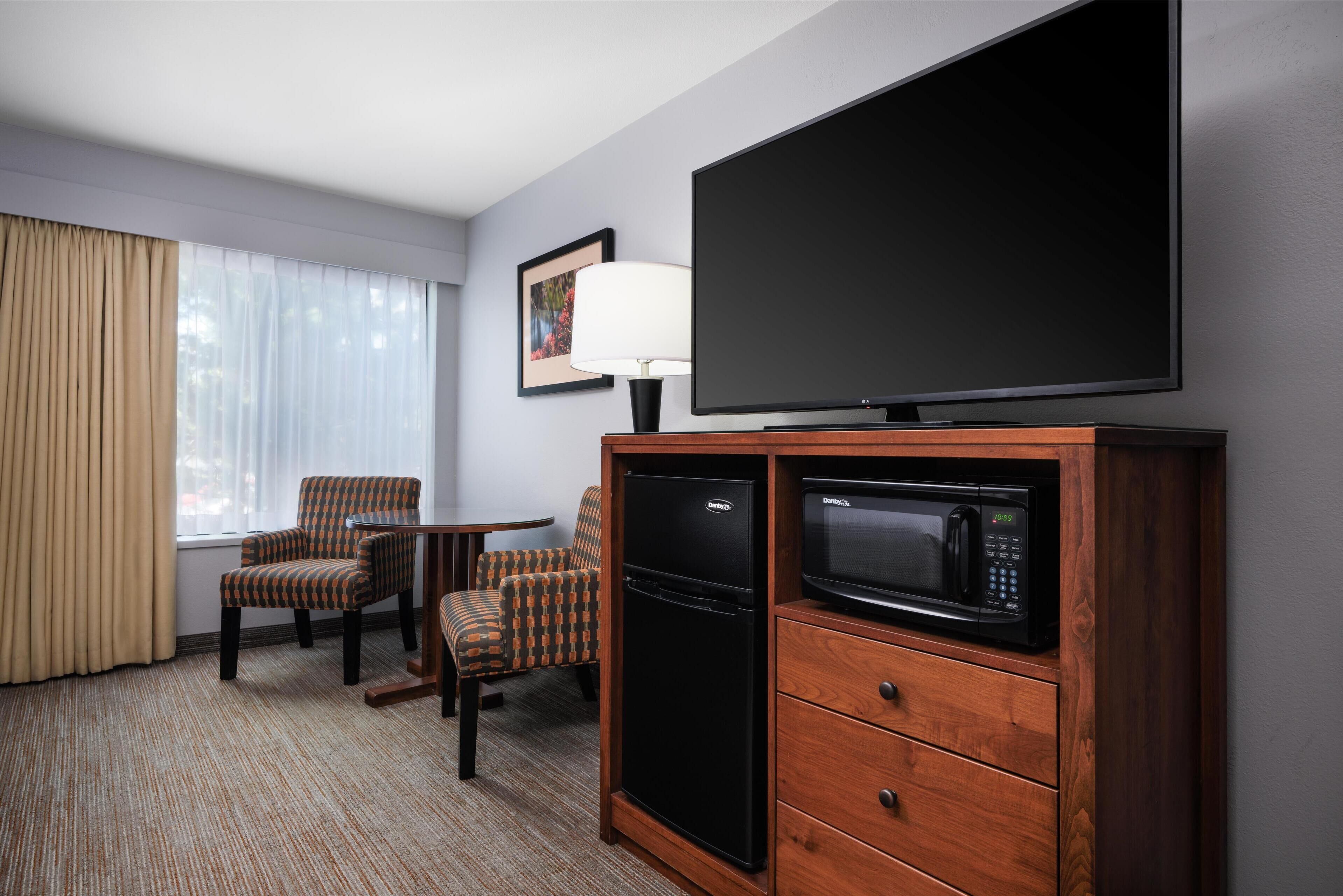 A hotel room with a flat-screen TV, a mini-fridge, a microwave, two chairs, a table, and a large window with curtains.