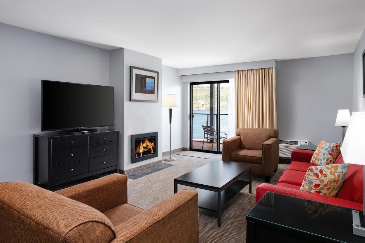 A modern hotel room with a flat-screen TV, a fireplace, a red sofa, a brown armchair, and a glass coffee table.