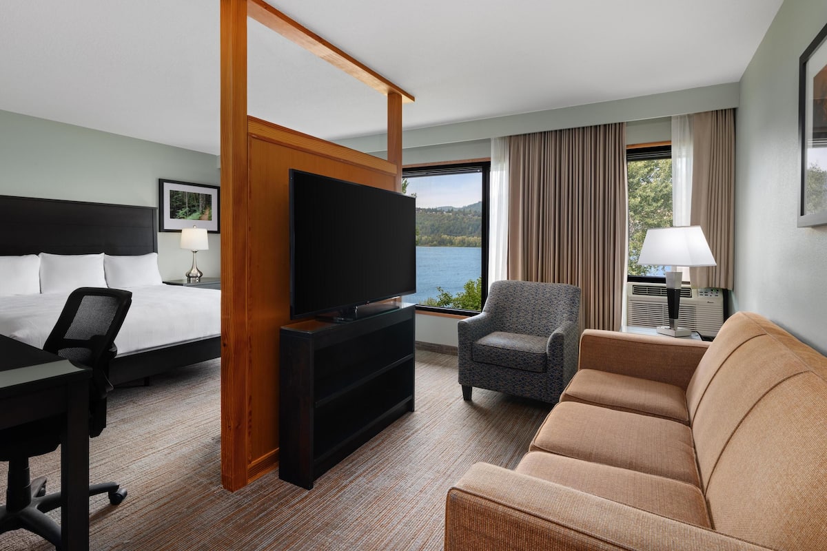 A hotel room with a bed, a TV, a desk, a chair, a sofa, and a large window with a view of a lake.