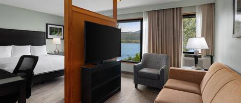 Une chambre d’hôtel équipée d’un lit, d’une télévision, d’un bureau, d’une chaise, d’un canapé et d’une grande fenêtre donnant sur un lac.