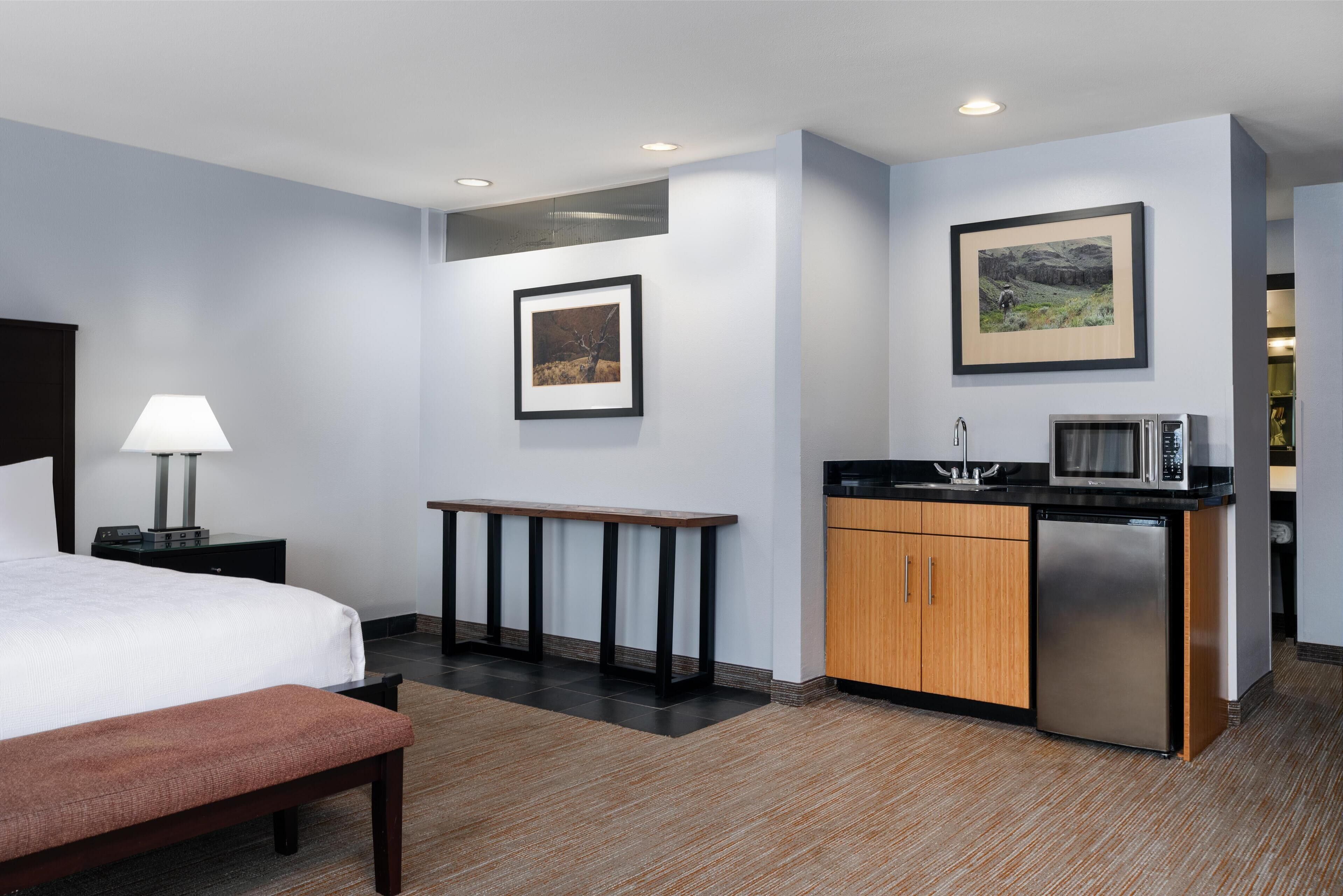 A hotel room with a bed, bedside table, lamp, a small kitchenette with a microwave, and a framed picture on the wall.