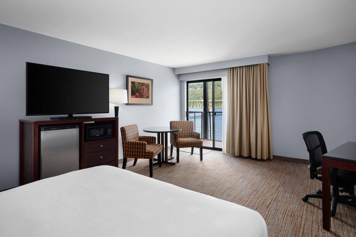 A hotel room with a bed, a desk with a flat-screen TV, a refrigerator, a microwave, two chairs, a small table, and a large window with curtains.